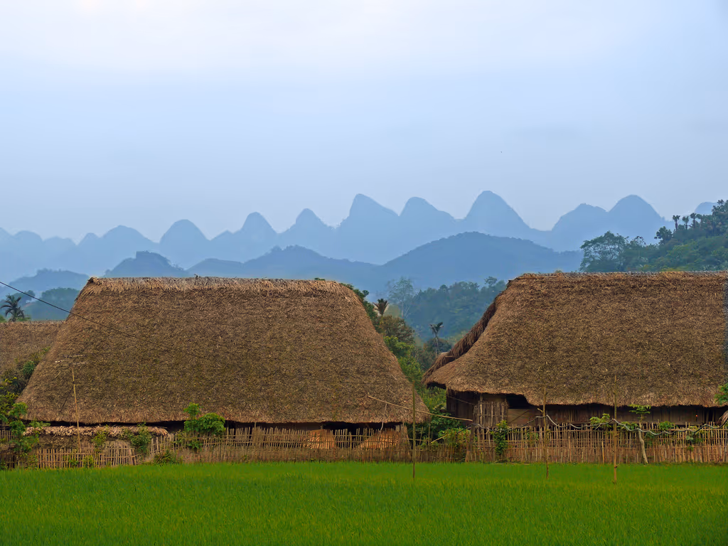 Thatched wooden houses in front of a row of pointed blue karst peaks near the road to Ban Gioc