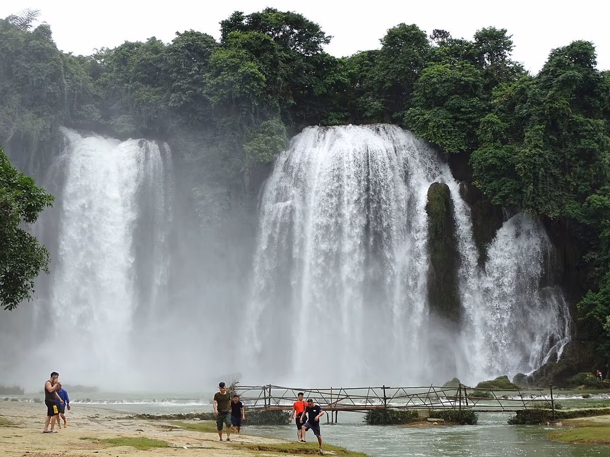 A wide curtain of white water on the Ban Gioc falls with trees above and visitors on a footbridge below