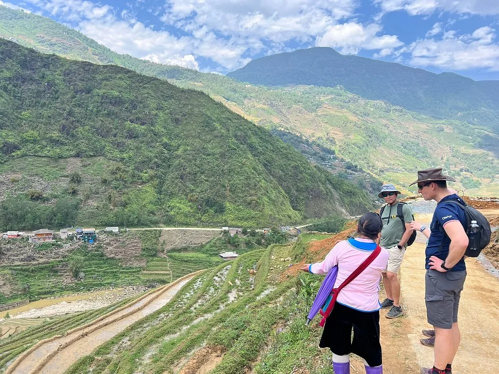 A local Black H'mong guide pointing out the Muong Hoa Valley to two Western trekkers under clear blue October skies near Lao Chai, Sapa