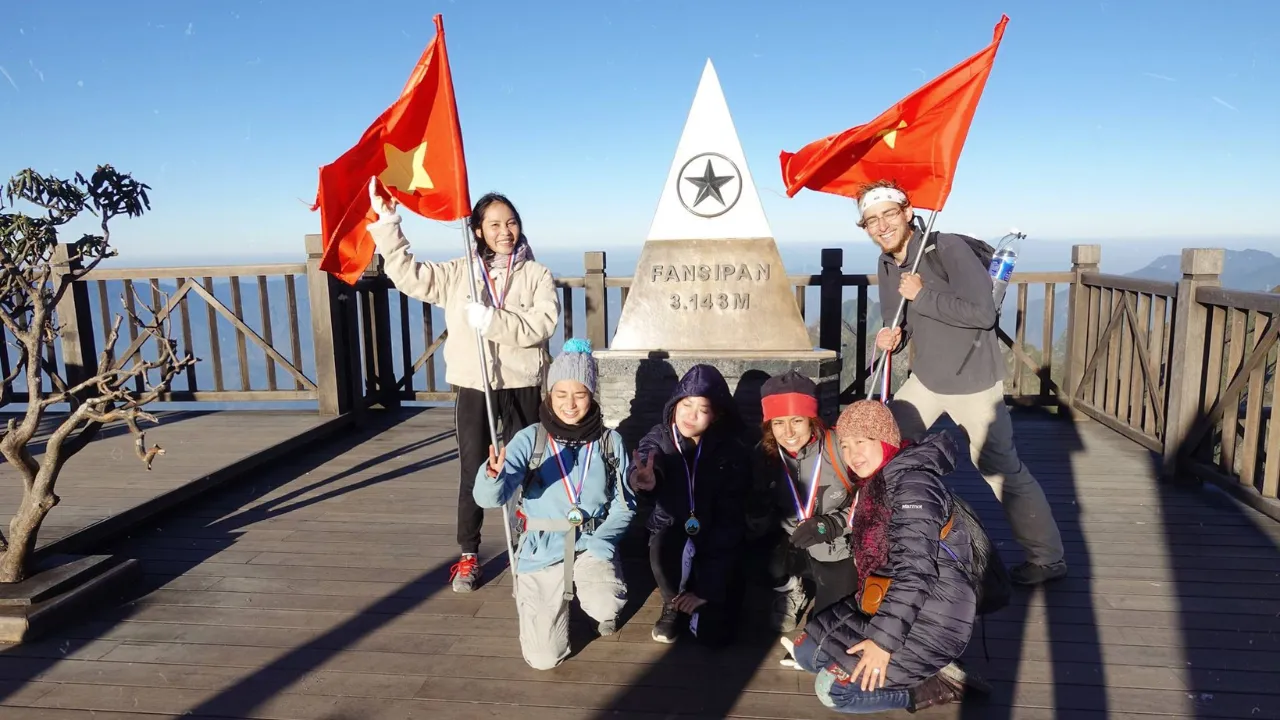 Group of trekkers celebrating at the Fansipan summit marker with Vietnamese flags after the one-day climb