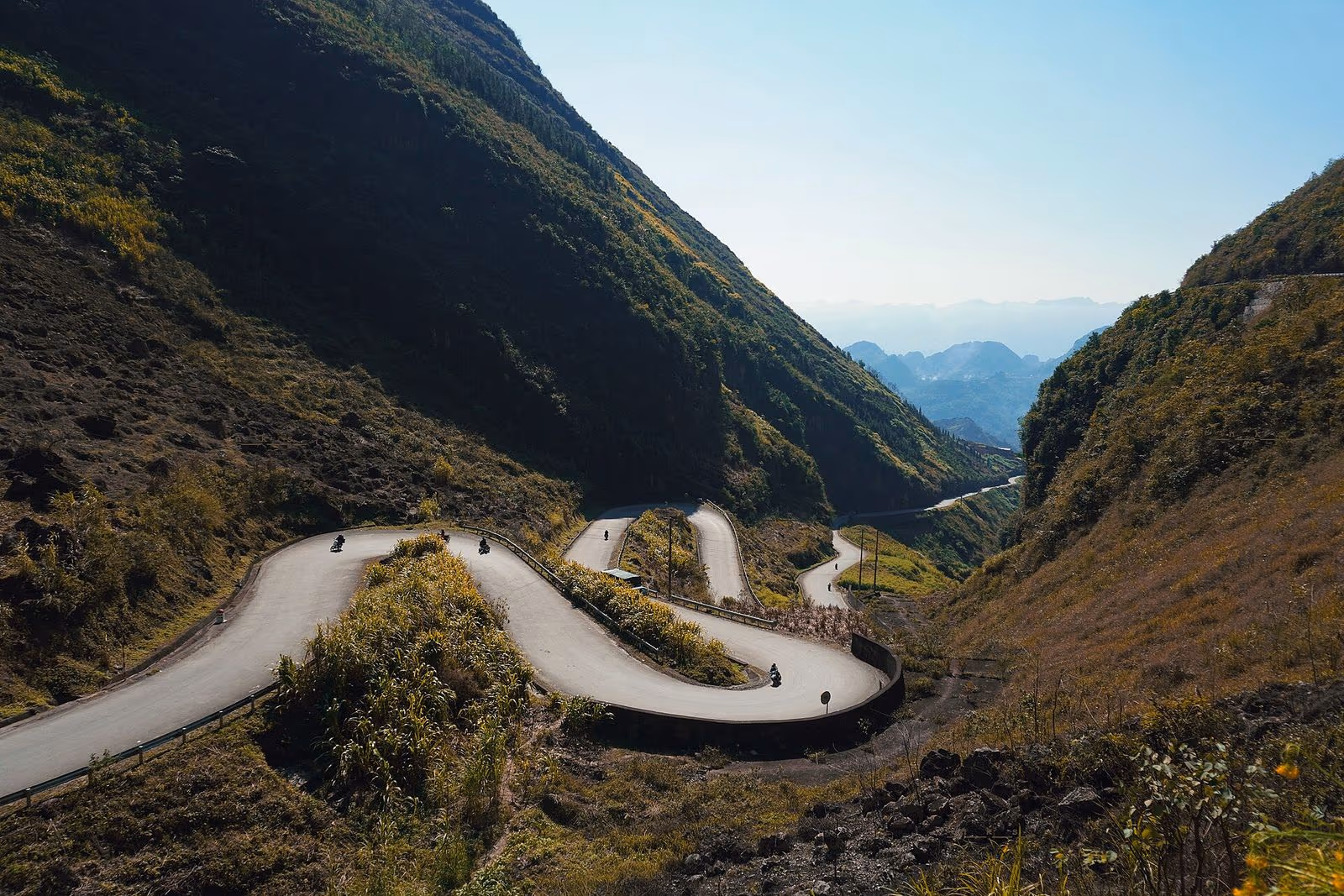 Hairpin bends of the Tham Ma slope road on the Dong Van plateau under clear blue sky, with a few motorbikes
