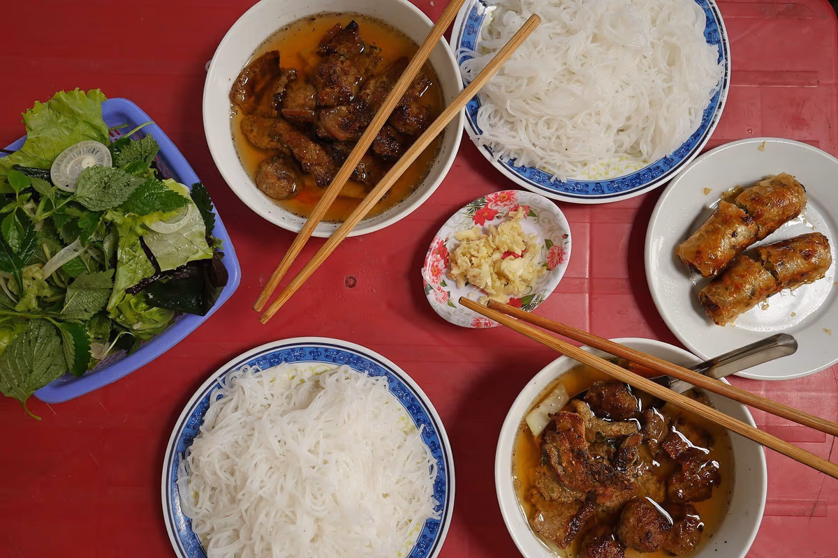 Top view of a bun cha meal on a red plastic table with bowls of grilled pork in broth, rice noodles, fresh herbs, garlic and fried spring rolls