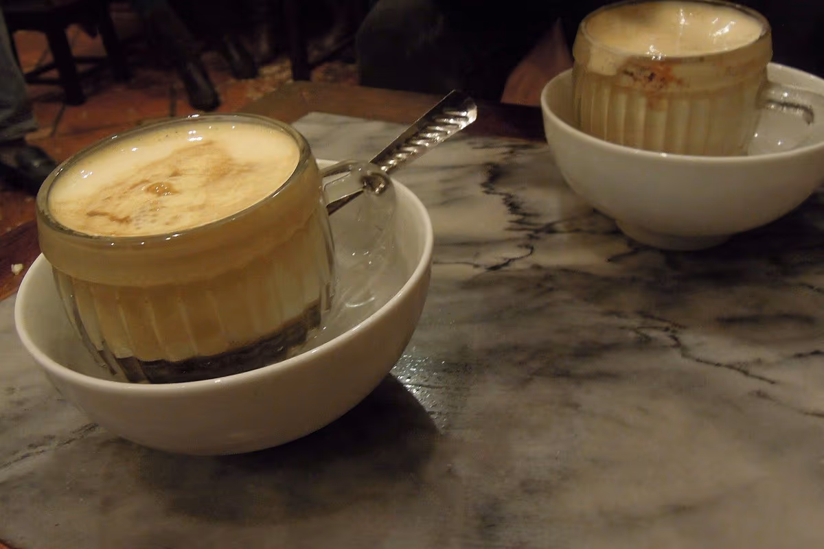 Two glass cups of egg coffee with a thick pale foam, each standing in a white bowl of warm water on a marble table