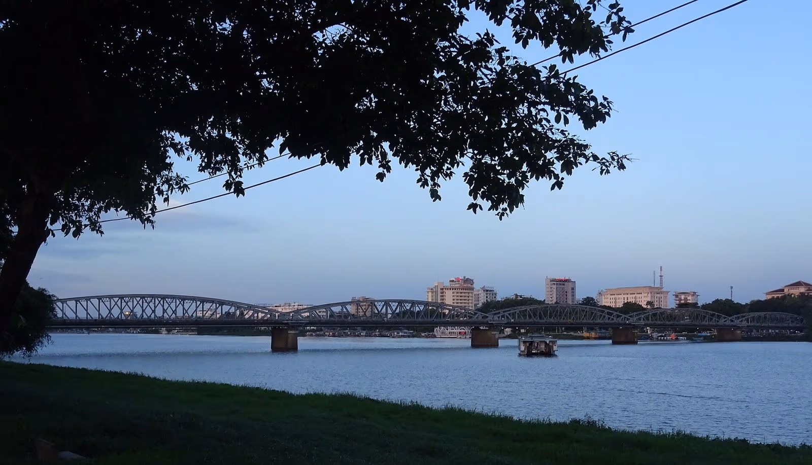 The long iron-arched Truong Tien Bridge across the Perfume River at dusk, with a small boat on the water and city buildings behind