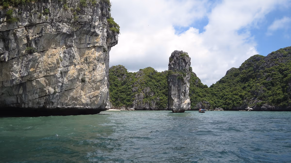 A narrow limestone stack standing in shallow water beside a pale cliff in Lan Ha Bay near Cat Ba Island