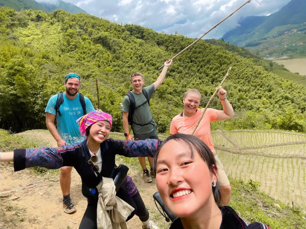 Guide Tzu Hang selfie with trekking group holding bamboo poles in Muong Hoa Valley, terraced rice fields visible in the background, Sapa, Vietnam