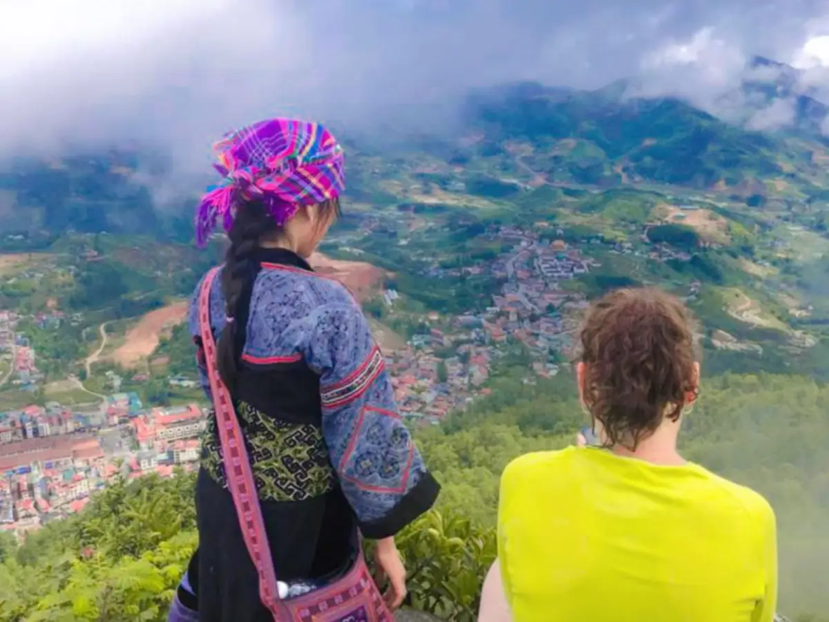 Black H'mong guide in traditional blue and pink embroidered outfit with colorful headdress, standing with traveler looking down at Sapa town from above