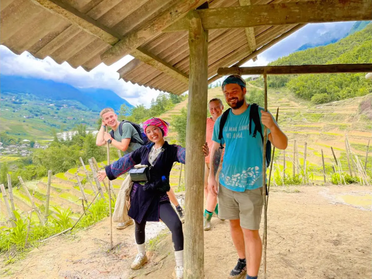 Black H'mong guide in traditional black and pink outfit with trekking group under a shelter, rice terraces in background, Ta Van Sapa Vietnam