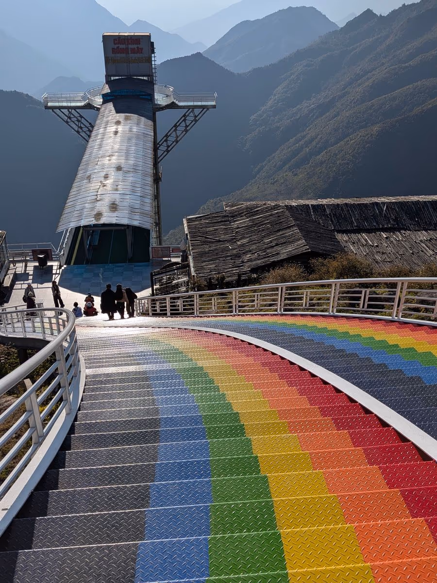 Rainbow-painted stairs leading down to the glass elevator tower at Rong May Glass Bridge, with the Hoang Lien Son mountains behind