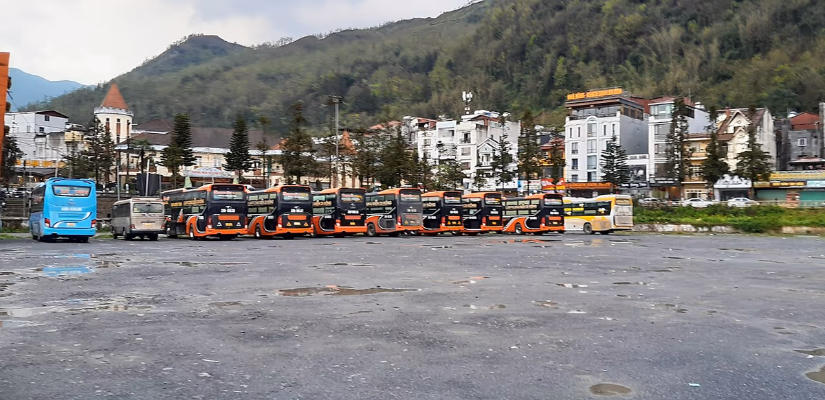 A row of orange and black sleeper buses parked in a large lot in Sapa town, with a church tower and hills behind
