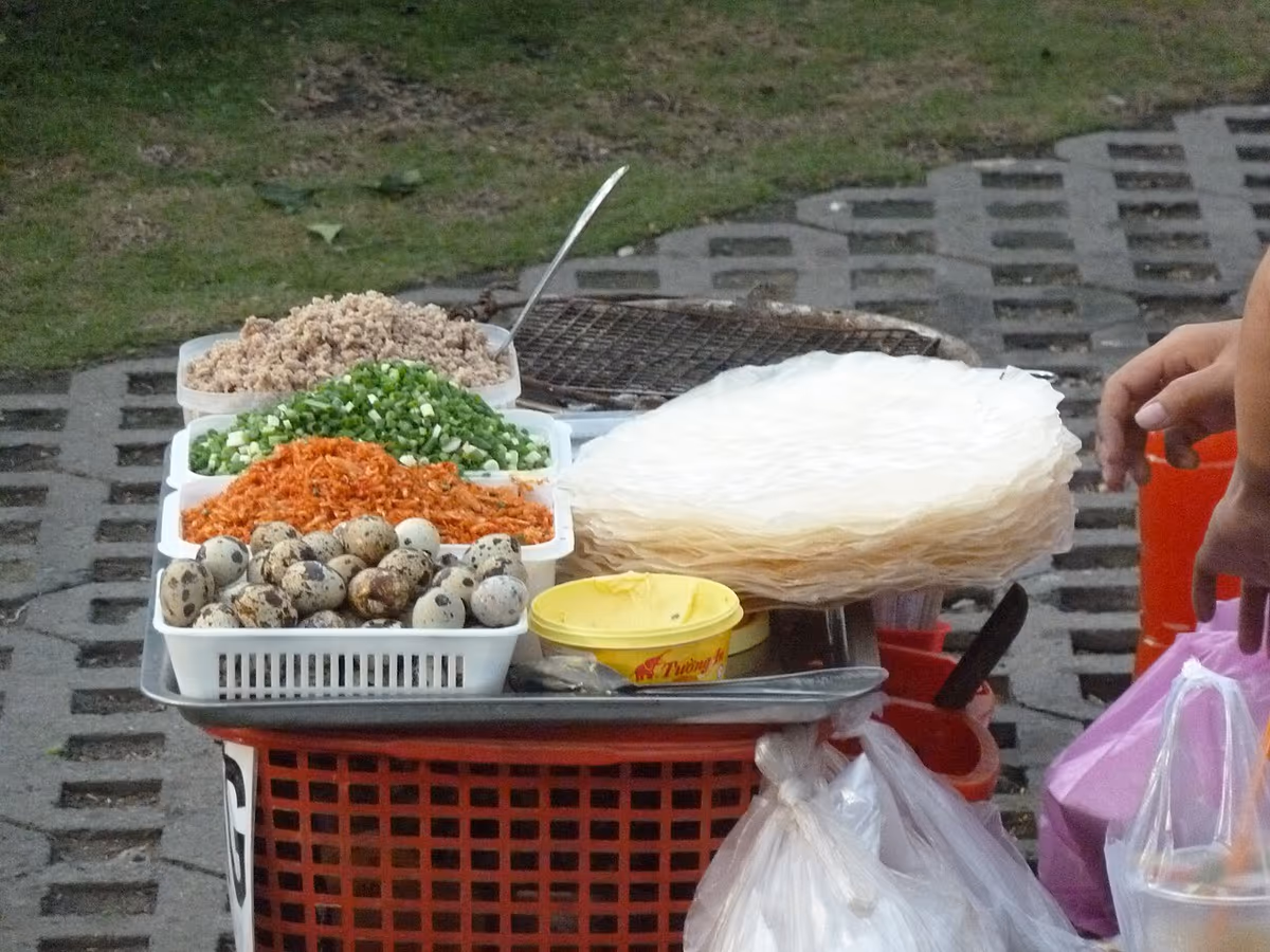 Street stall tray with quail eggs, minced pork, green onions, dried shrimp and a stack of rice paper beside a charcoal grill