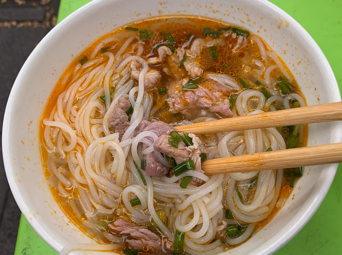 Bowl of bun bo Hue with beef slices, rice noodles and spring onion in a red-orange broth, chopsticks resting in the bowl