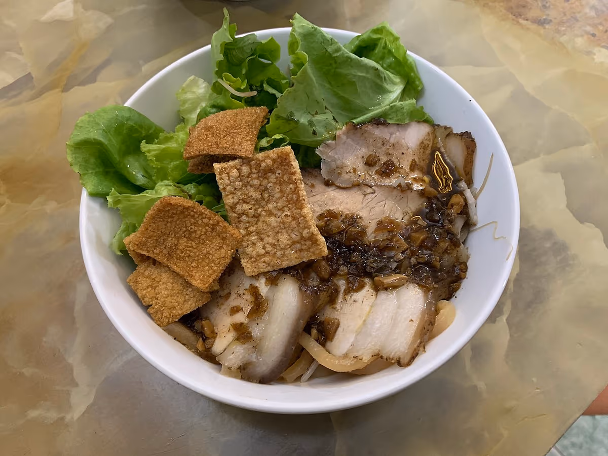 White bowl of cao lau with slices of roast pork, crisp fried noodle squares, lettuce and sauce