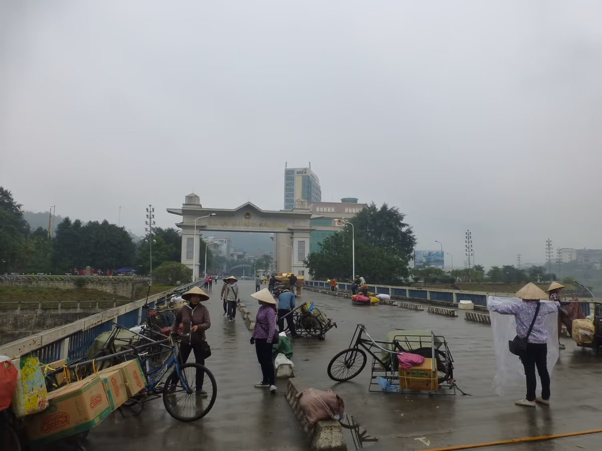 The Vietnamese border gate arch at the end of the Lao Cai bridge, with porters in conical hats and handcarts on a wet road